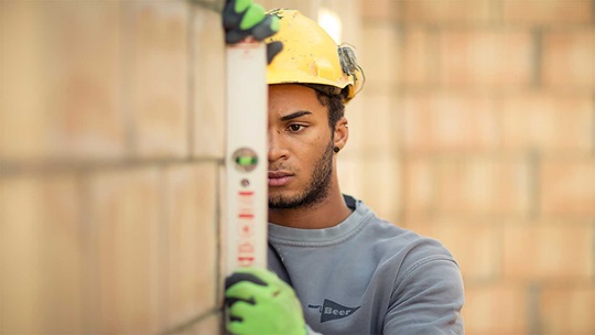 Ein Maurer steht mit Wasserwage bei einer Ziegelsteinmauer