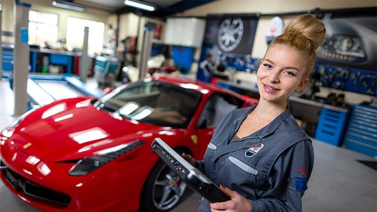 Eine Automechatronikerin steht vor einem roten Sportwagen und hält ein Notebook in der Hand