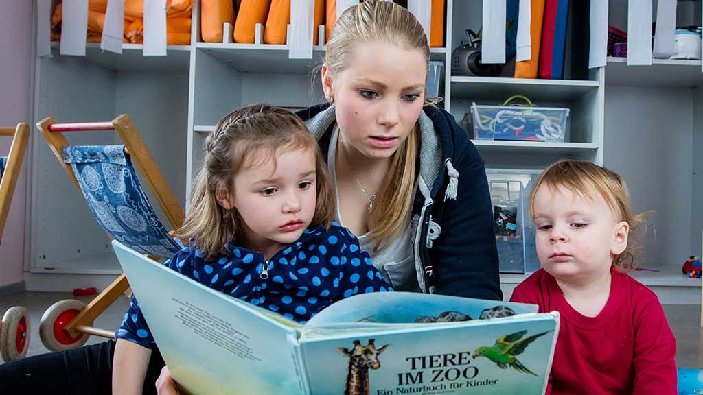 Eine Fachfrau Betreuung sitzt mit zwei Kindern am Boden und schaut mit ihnen ein Buch über Tiere im Zoo an