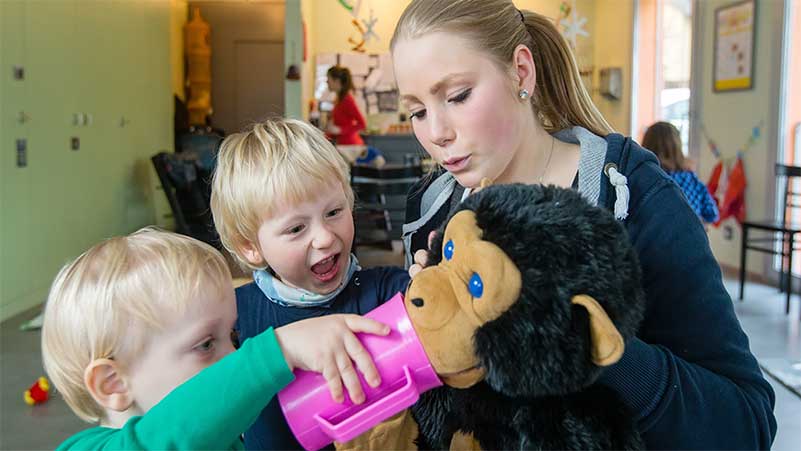 Eine Fachfrau Betreuung hält einen Teddy-Affen in der Hand und zwei kleine Kinder wollen ihm mit einem pinken Becher etwas zu trinken geben