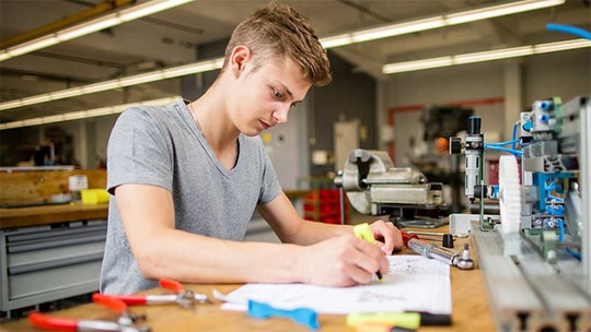 Ein Polymechaniker sitzt an seinem Pult und studiert einen Plan auf Papier