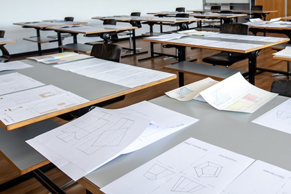 Auf den Tischen eines leeren Klassenzimmers liegen Unterlagen.